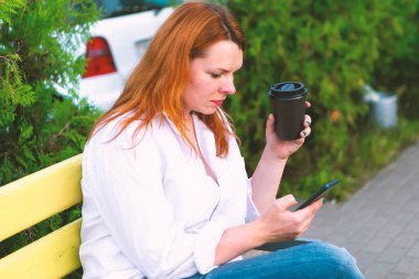 Elinde telefon ve kahve fincanı olan kız parkta bankta oturuyor, kapat.