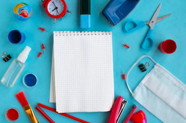 Mockup notebook, school supplies and medical mask on blue background, top view. Back to school concept, school quarantine concept