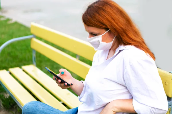 Woman in medical mask with phone sits on the bench in the park, coronavirus concept