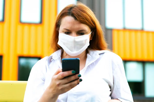 Woman in medical mask with phone sits on the bench in the park, coronavirus concept