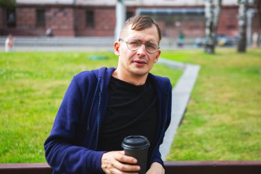 Bankta oturan genç adam yaz parkında kahve içiyor.