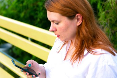 Cep telefonlu bir kadın parktaki bankta oturuyor, açık hava yaz arkaplanı.