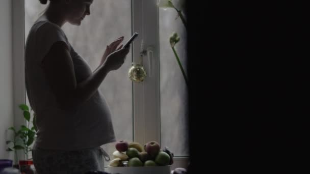 Веселая беременная женщина с мобильным телефоном на кухне — стоковое видео