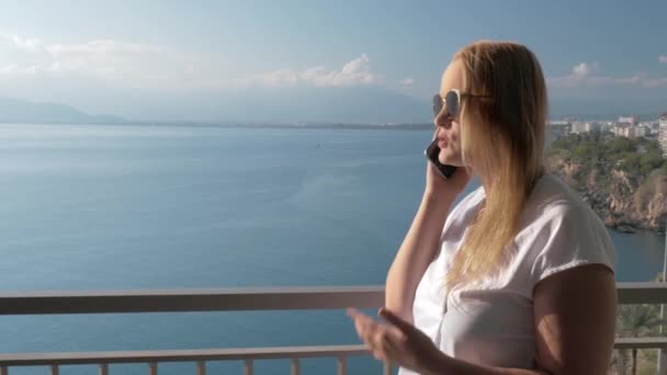 Una mujer de pelo rubio hablando con un teléfono en el balcón de un hotel cerca del paisaje marino — Vídeo de stock