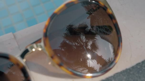 Close-up shot of female sunglasses on swimming pool border with palm and sun reflection. Vacation time