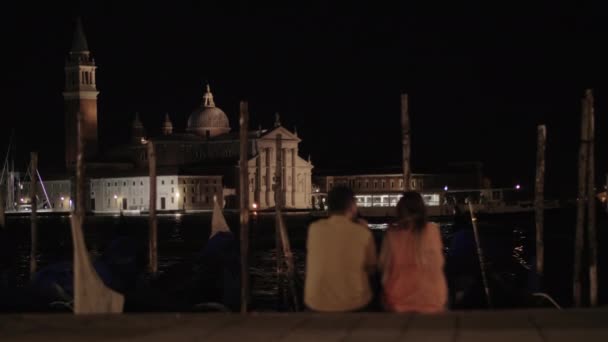 Una pareja sentada en el terraplén frente a la vista nocturna de Venecia — Vídeos de Stock