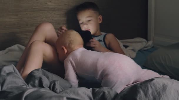 Un chico con una tableta y su hermanita en una cama desordenada — Vídeo de stock