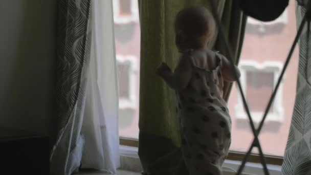 En baby flicka i en övergripande leka med gardiner nära det stora fönstret — Stockvideo