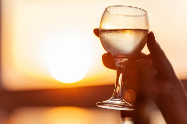 A misted glass of cold white wine in a womans hand, as gold as sunset in the background — Stock Photo, Image