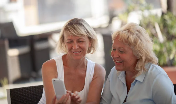 Dos mujeres de mediana edad sentadas afuera y sonriendo mientras miran la pantalla de un teléfono inteligente —  Fotos de Stock