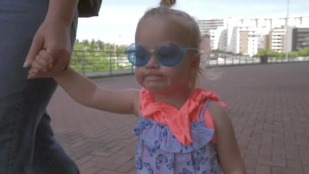 Uma menina de cabelos claros em um azul muito elegante rodada óculos de sol andando segurando as mães mão — Vídeo de Stock