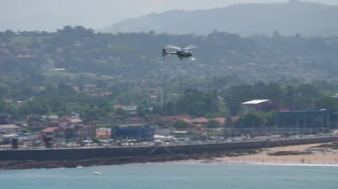 Askeri helikopter kıyı kenti boyunca ağaçların ve binaların üzerinde alçaktan uçuyor.