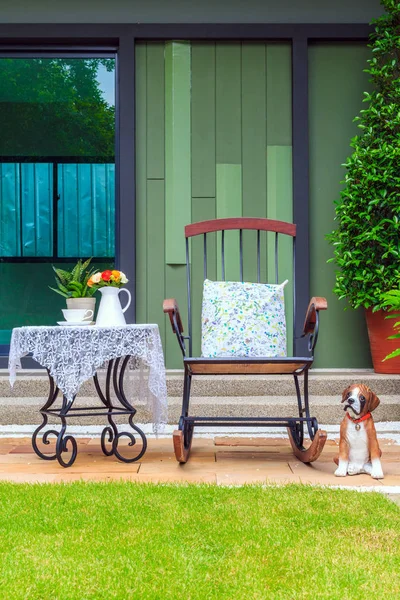 A tea set and flowers in a white pot on the vintage table with and wooden rocking chair with a dog doll in the garden.
