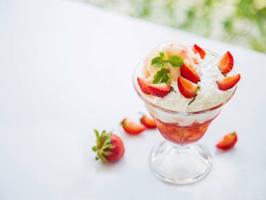Yan taraftaki manzarada öğleden sonra doğanın arka planında cam pencerenin yanındaki beyaz bir masanın üzerinde nane yapraklı lezzetli çilekli dondurma var..