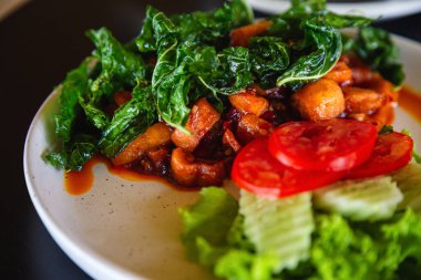 Yağda kızartılmış domuz eti ve köri ezmesini kapayın. Beyaz bir tabakta domates ve salatalık ile siyah bir masaya süslenmiş fesleğen, Tayland yemeği kavramı.