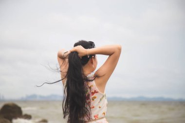 Güzel elbiseli genç bir Asyalı kadının arka portresi kapalıyken kasvetli bir günde kuyruğunu denizin önüne kıstırıyor.