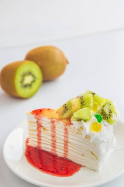 Yakından bakınca, kivi meyveleri bulanık arka planda beyaz bir masada beyaz bir tabakta çilek soslu lezzetli bir kivili krep görüyorum..