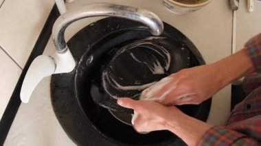 Eller siyah bir tabağı sünger ve bulaşık deterjanıyla yıkıyor, köpük yapıyor ve durulanıyor.