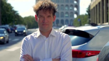 Beyaz tişörtlü, gülümseyen adam şehirdeki kameraya bakıyor.