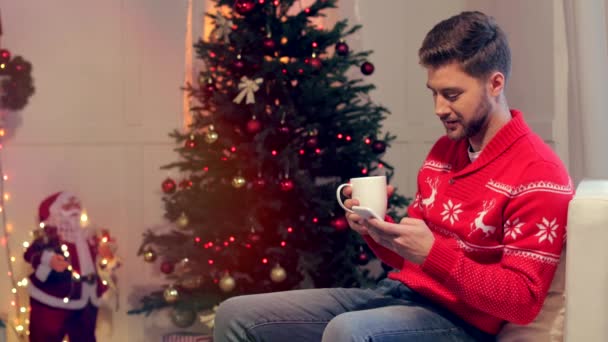 快乐的年轻人坐在沙发上与咖啡和使用智能手机在圣诞节装饰客厅 — 图库视频影像
