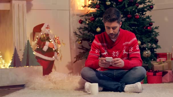 Jovem Atraente Sentado Chão Usando Smartphone Sala Estar Decorada Natal — Vídeo de Stock