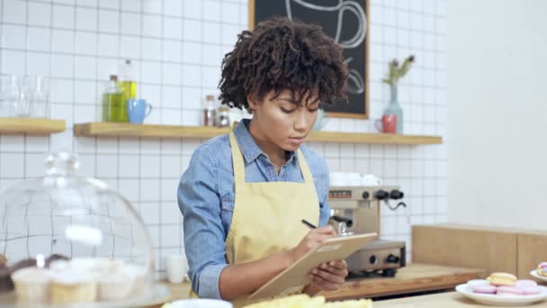 カフェでカウンターの後ろにクリップボードに書く美しいアフリカ系アメリカ人の女性レジ係 — ストック動画
