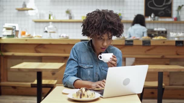 美丽的非洲裔美国女性自由职业者坐在桌子上 喝咖啡 微笑着 在咖啡馆使用笔记本电脑 — 图库视频影像