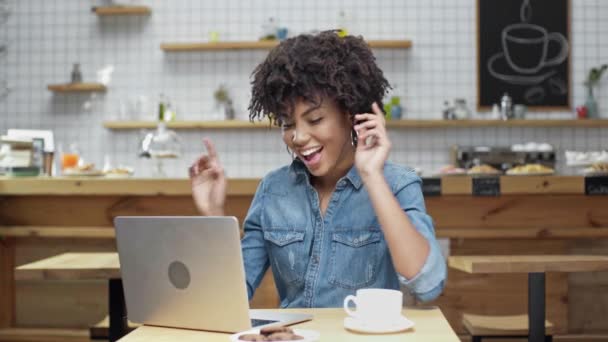 ラップトップを使用して カフェで音楽を聴くイヤホンで美しい幸せなアフリカ系アメリカ人女性フリーランサー — ストック動画