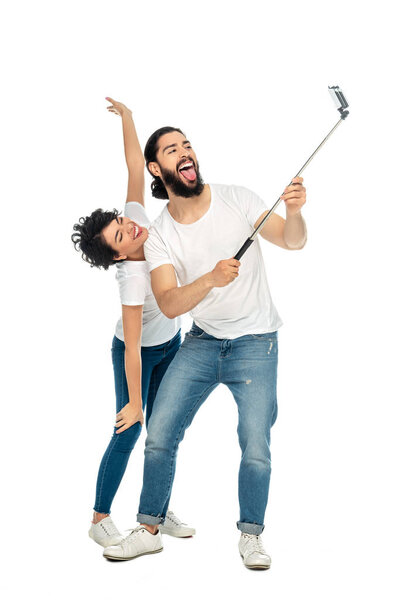 happy latin man showing tongue near brunette woman with outstretched hands while holding selfie stick and taking selfie isolated on white 