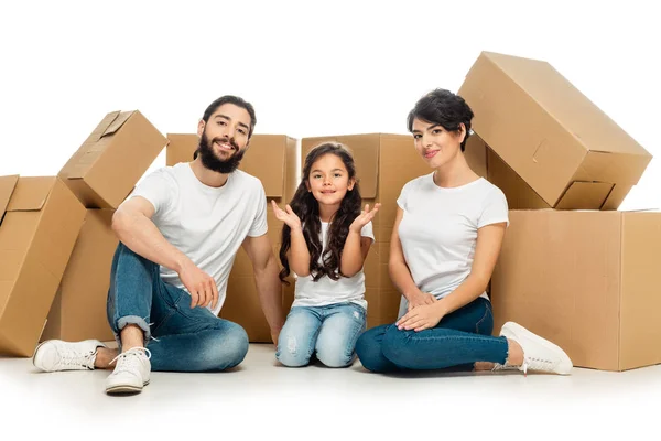 cheerful latin parents sitting near cute daughter and carton boxes isolated on white