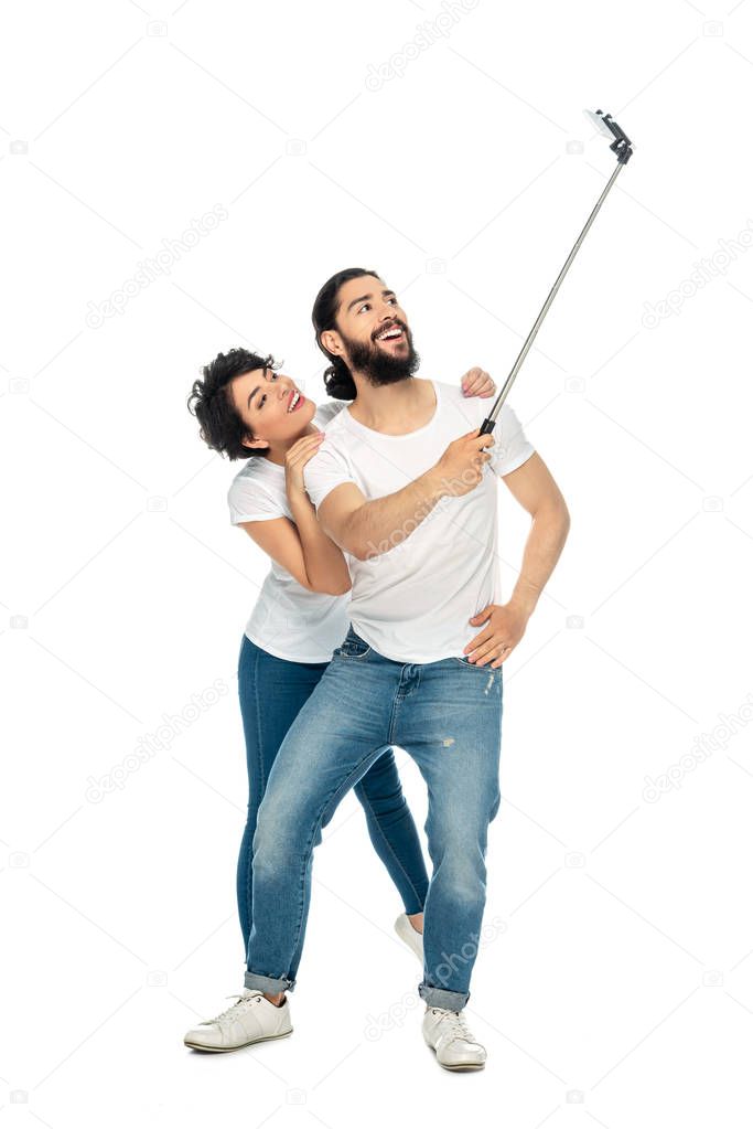 happy latin man smiling near brunette woman while holding selfie stick and taking selfie isolated on white 