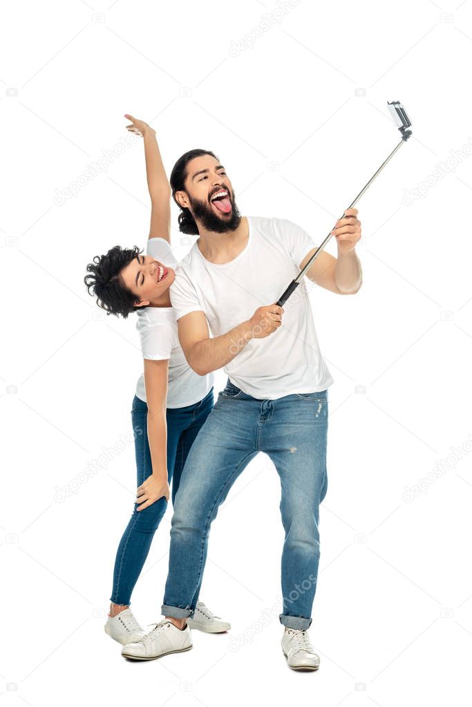 happy latin man showing tongue near brunette woman with outstretched hands while holding selfie stick and taking selfie isolated on white 