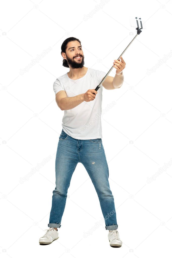 happy latin man holding selfie stick and taking selfie while standing isolated on white 
