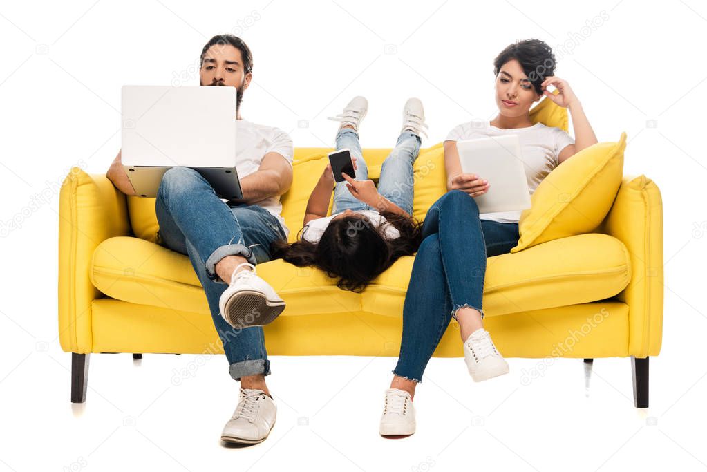 kid lying on sofa and holding smartphone with blank screen near latin parents using gadgets isolated on white 