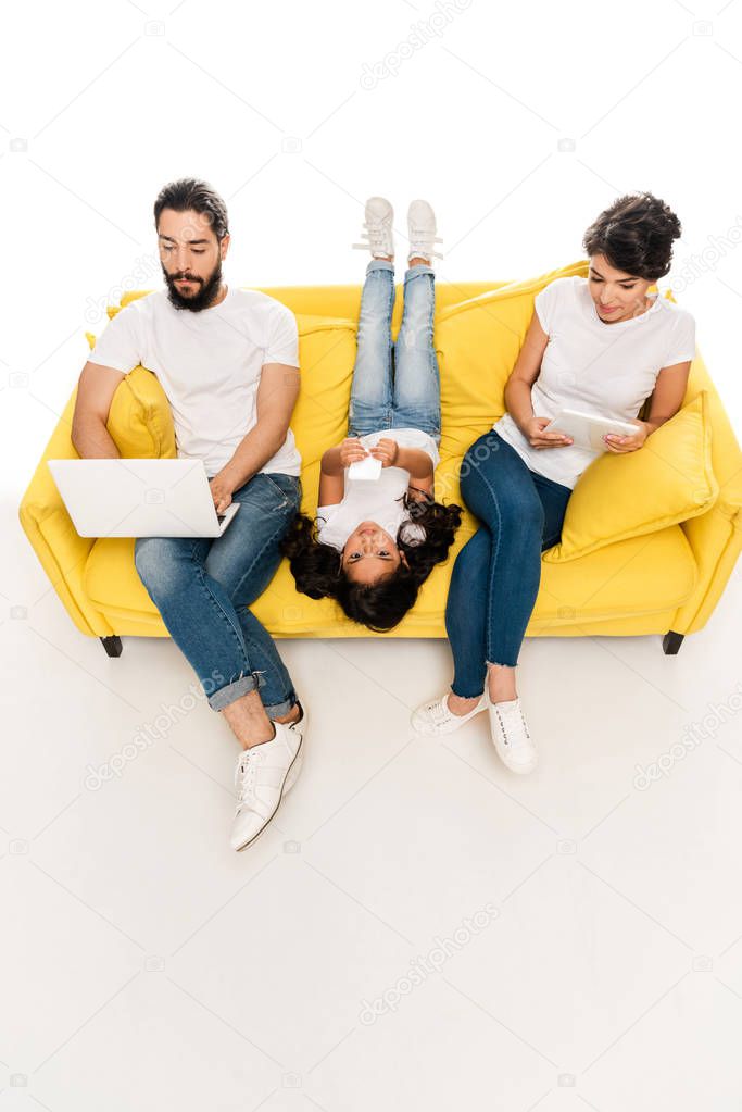 overhead view of kid lying on sofa and holding smartphone near latin parents using gadgets isolated on white  