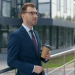 Selective focus of businessman drinking coffee to go, smiling and looking at camera near building