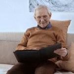 Cheerful pensioner in glasses smiling while looking at photo album