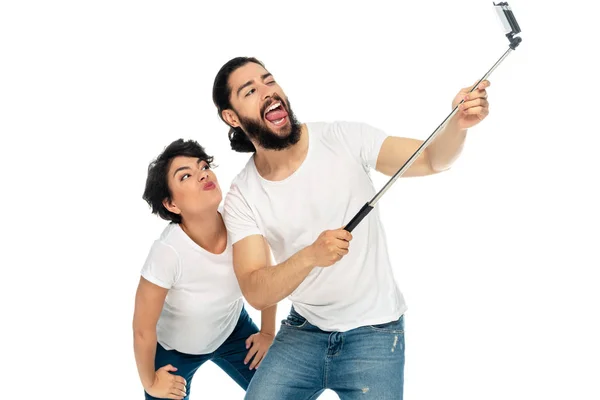 Felice uomo latino mostrando la lingua vicino alla donna bruna e tenendo bastone selfie mentre prende selfie isolato su bianco — Foto stock