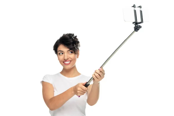 Enfoque selectivo de la mujer latina feliz sosteniendo palo selfie y tomando selfie aislado en blanco - foto de stock