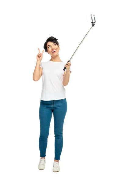 Alegre mujer latina mostrando signo de paz mientras sostiene palo selfie y tomar selfie aislado en blanco - foto de stock