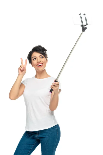 Mujer latina feliz mostrando señal de paz mientras sostiene el palo selfie y toma selfie aislado en blanco - foto de stock
