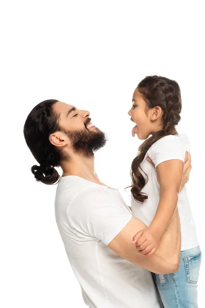 Feliz padre latino sonriendo mientras sostiene en brazos linda hija mostrando la lengua aislada en blanco - foto de stock