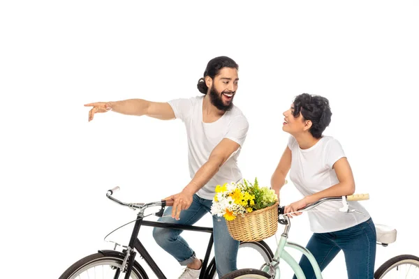 Hombre latino feliz señalando con el dedo mientras monta bicicleta cerca de mujer atractiva aislado en blanco - foto de stock