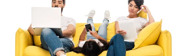Plan panoramique d'un enfant allongé sur un canapé et tenant un smartphone avec écran vide près de parents latins à l'aide de gadgets isolés sur du blanc — Photo de stock