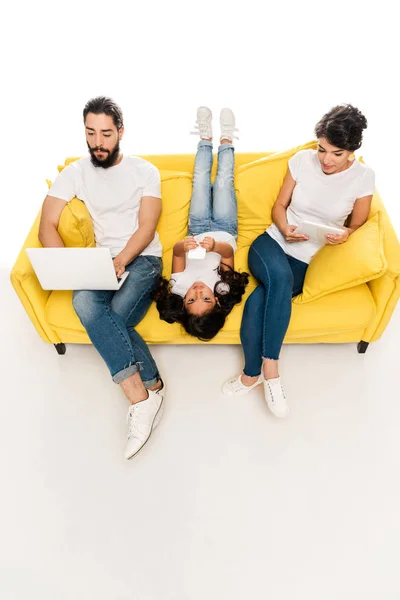 Vista aérea del niño acostado en el sofá y sosteniendo el teléfono inteligente cerca de los padres latinos utilizando gadgets aislados en blanco - foto de stock