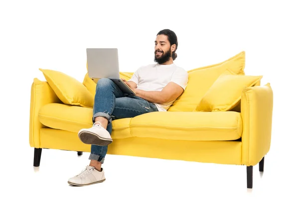 Glücklicher bärtiger lateinischer Mann in blauen Jeans sitzt auf gelbem Sofa und benutzt Laptop isoliert auf weißem Hintergrund — Stockfoto