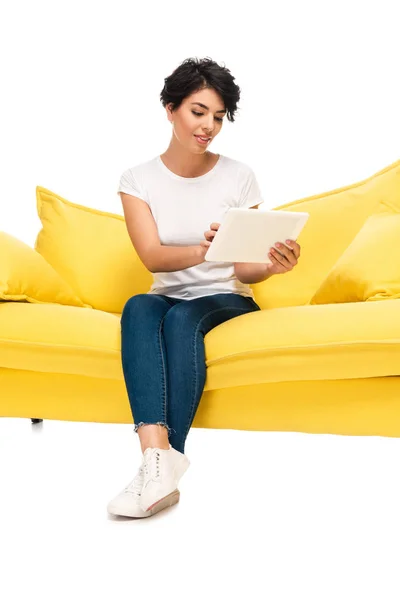 Mulher latina feliz usando comprimido digital enquanto sentado no sofá isolado no branco — Fotografia de Stock