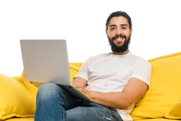 Uomo latino sorridente utilizzando il computer portatile e seduto sul divano isolato su bianco — Foto stock