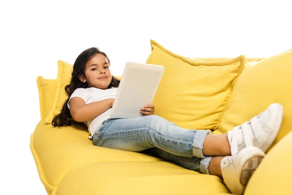 Селективный фокус счастливого латинского ребенка с помощью цифрового планшета, лежащего на диване, изолированном на белом — стоковое фото
