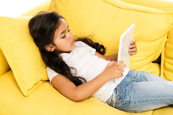 Vista aérea de un lindo niño latino acostado en un sofá amarillo y usando una tableta digital aislada en blanco - foto de stock